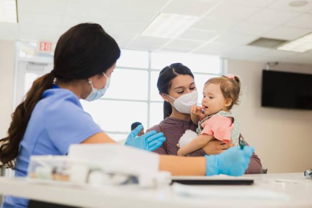 Volunteer nurse cares for baby at free clinic A female healthcare professional gestures as she talks to a young mother and the woman's baby. The nurse and mother are wearing protective face masks during the coronavirus outbreak. family nurse practitioner stock pictures, royalty-free photos & images